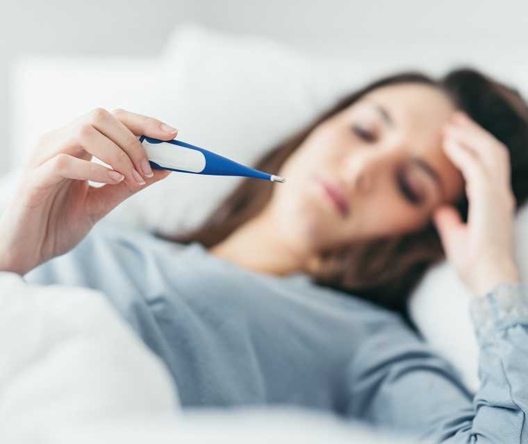 Eine Frau liegt im Bett und schaut auf ein Fieberthermometer